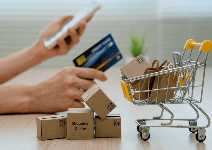 Person holding a smartphone and a credit card near a miniature shopping cart filled with boxes labeled “Shopping Online, ”representing e-commerce activity and the convenience of digital marketplaces.